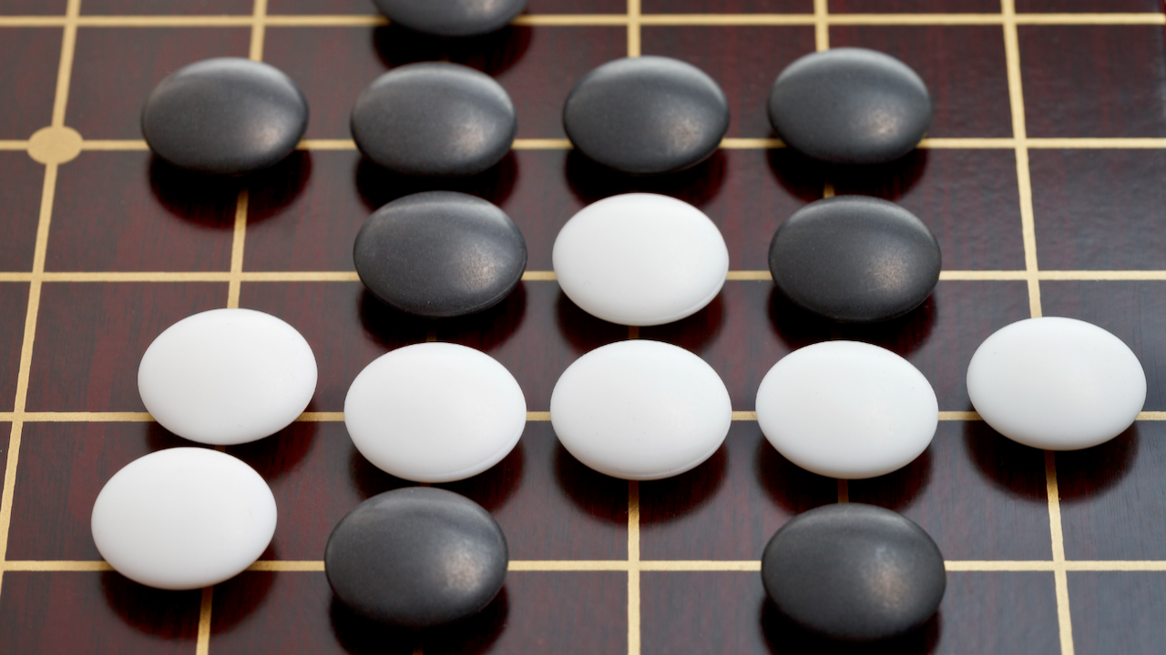Black and white stones are arranged on a wooden Go board with a grid pattern. The stones are grouped in a way that suggests part of a Go game is in progress.
