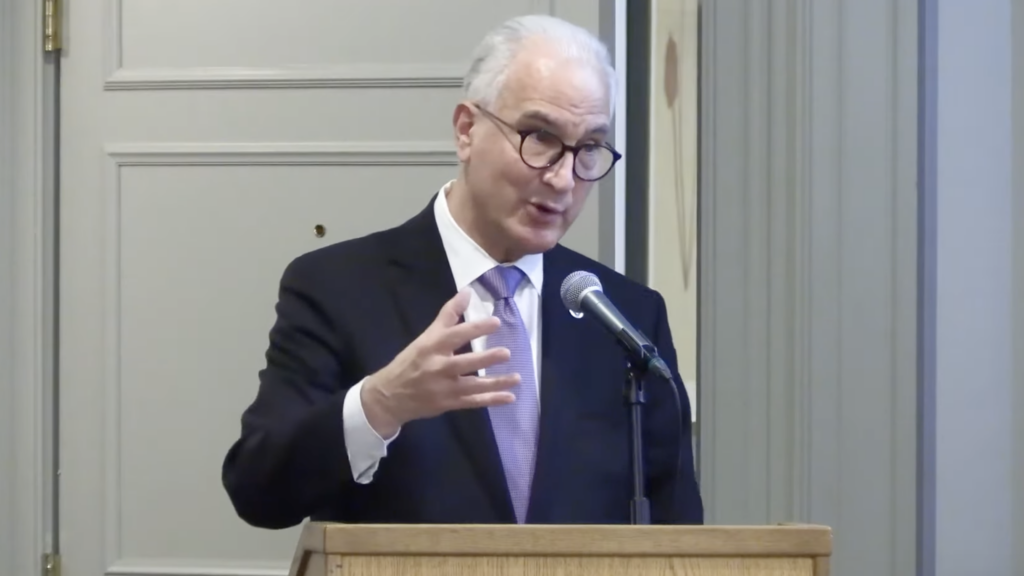 A man with gray hair and glasses, wearing a dark suit and light purple tie, speaks at a podium with a microphone, gesturing with his left hand in a formal indoor setting.