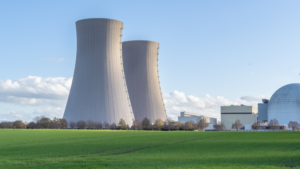 Two large cooling towers and several industrial buildings of a nuclear power plant stand next to a green grassy field under a blue sky with some clouds.
