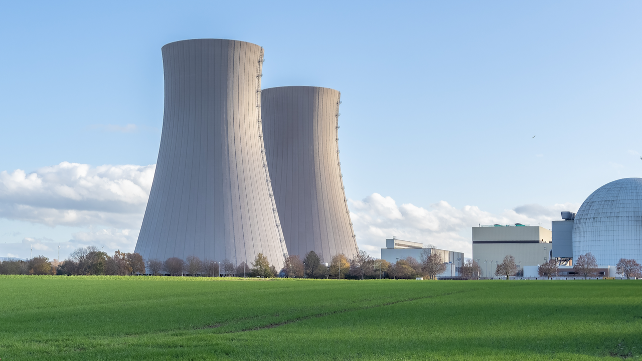 Two large cooling towers and several industrial buildings of a nuclear power plant stand next to a green grassy field under a blue sky with some clouds.