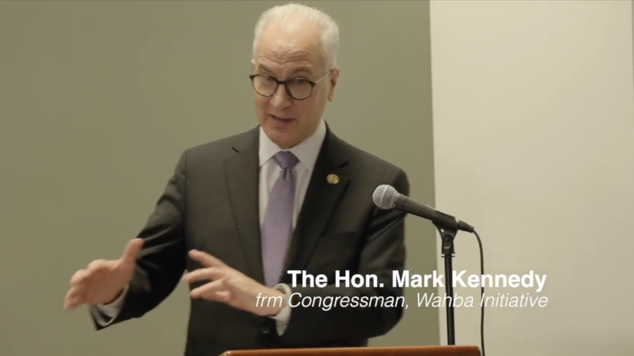 A man in a suit and purple tie speaks at a podium with a microphone. Text on the image reads: The Hon. Mark Kennedy, frm Congressman, Wahba Initiative. He gestures with his hands while speaking.