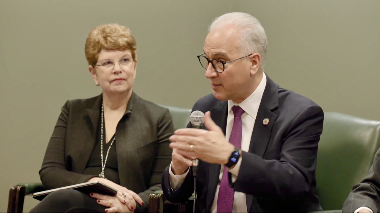A man in a suit and glasses speaks into a microphone, gesturing with his hand, while a woman with short red hair listens attentively, holding a notebook in her lap.