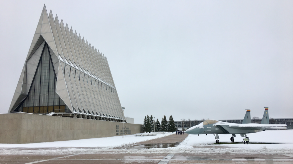 A modern, triangular-roofed chapel stands beside a snow-covered courtyard with a fighter jet display at the U.S. Air Force Academy on a cloudy winter day.