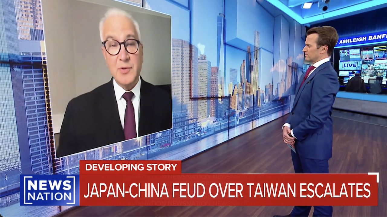 A news anchor stands in a studio, listening to a man in a suit and glasses speaking via video call. On-screen text reads: DEVELOPING STORY: JAPAN-CHINA FEUD OVER TAIWAN ESCALATES.