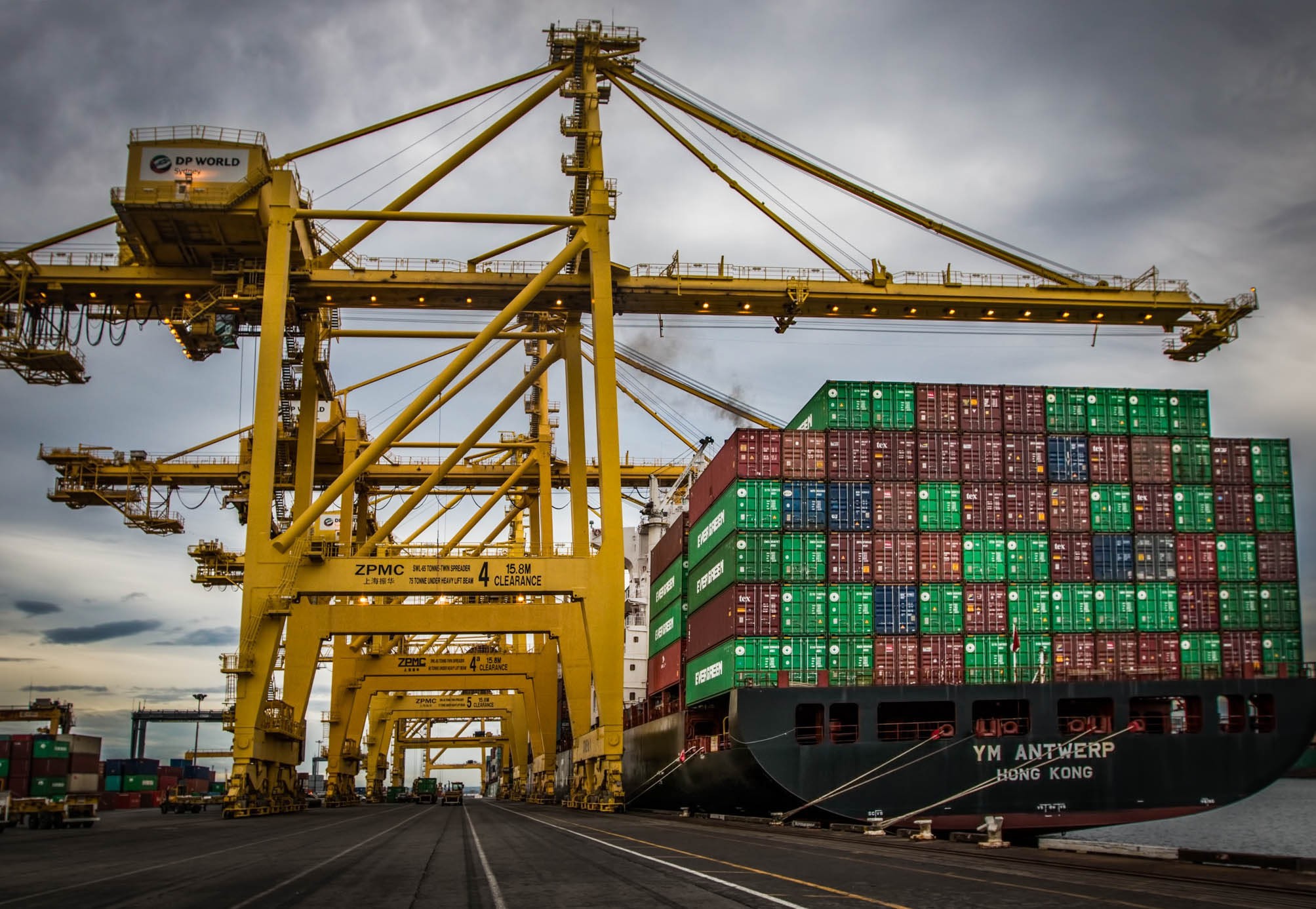 A large container ship loaded with colorful shipping containers is docked at a port, with tall yellow cranes lifting containers on and off the ship under a cloudy sky.