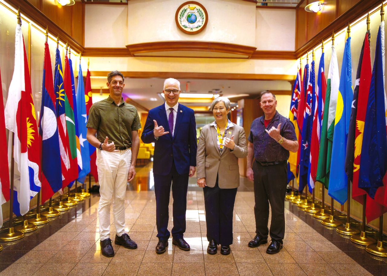 Four people stand smiling and posing indoors between two rows of international flags, with a circular emblem on the wall behind them. Three make a shaka hand gesture. The floor is shiny and reflective.