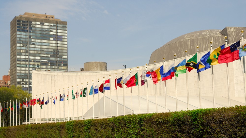 Colorful international flags line the front of the United Nations Headquarters in New York City, with modern high-rise buildings and a clear blue sky in the background.