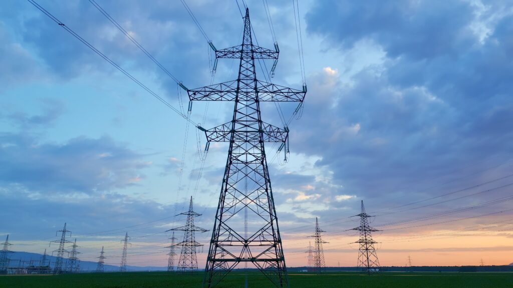 A row of tall electrical transmission towers stands on a green field under a cloudy sky at sunset, carrying power lines across the landscape.