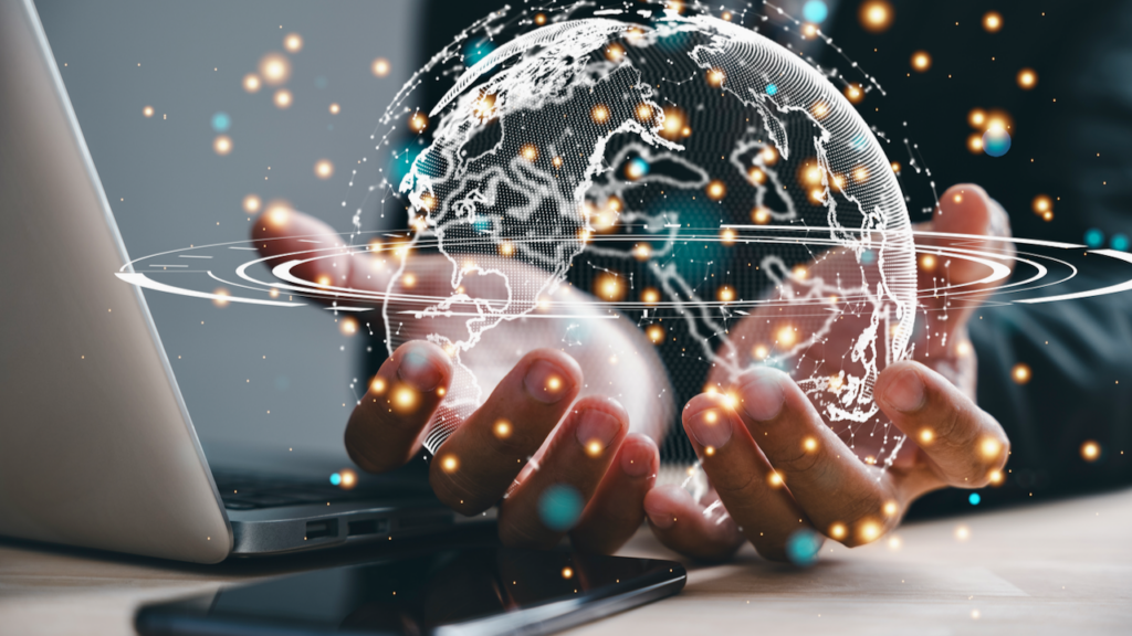A person’s hands are open beside a laptop and smartphone, with a glowing digital globe and light particles floating above, symbolizing global technology and connectivity.