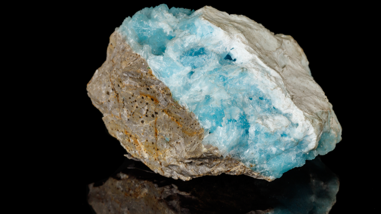 A rough mineral specimen with pale blue crystals embedded in a matrix of tan and white rock, displayed against a black background.