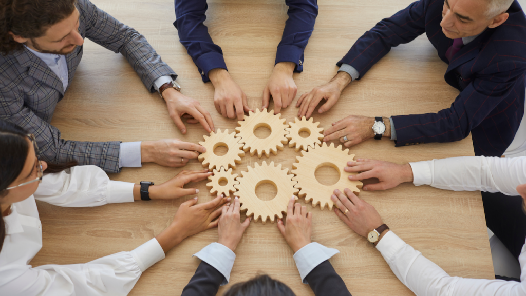 A group of people in business attire sitting around a table, each placing a hand on interlocking wooden gears, symbolizing teamwork and collaboration.